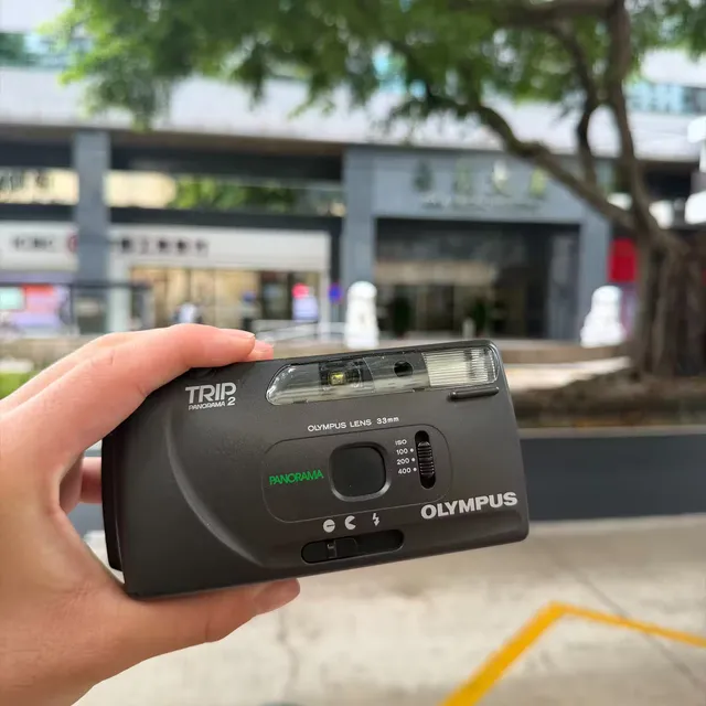 An Olympus Trip Panorama 2 compact camera held up in one hand on a street in Macau
